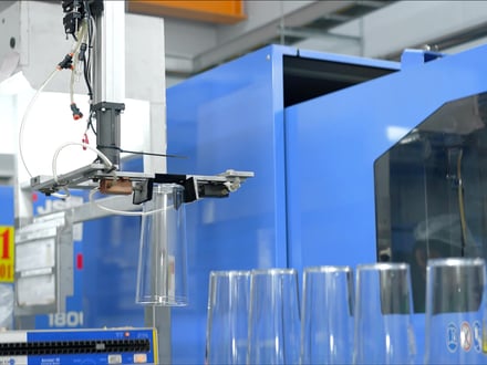 JSW 成形機から取り出した透明成形部品を払い出す AMP のロボット。民生用または食品飲料用途向けの光学透明材料で成形されている。