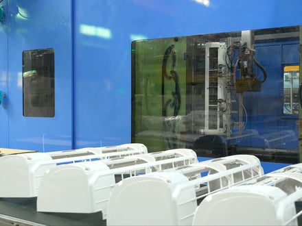 成形機の脇に並べられた AMP の家電筐体成形品。安全ガラス越しに金型とロボット取り出し装置が見える。